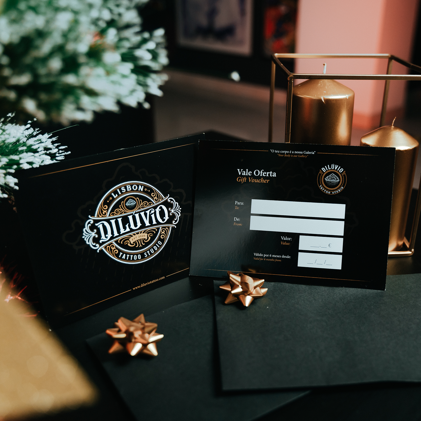 Gift voucher do Dilúvio Tattoo Studio exposto em ambiente festivo com laço dourado, envelope preto e velas decorativas, ideal para oferta de tatuagem em Lisboa