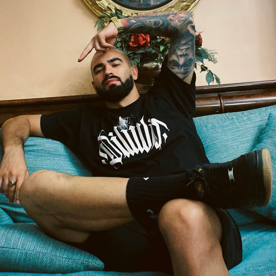 Pessoa sentada numa sala com decoração clássica, vestindo t-shirt oficial do merchandise do Dilúvio Tattoo Studio. Estilo casual com destaque para tatuagens e moda streetwear