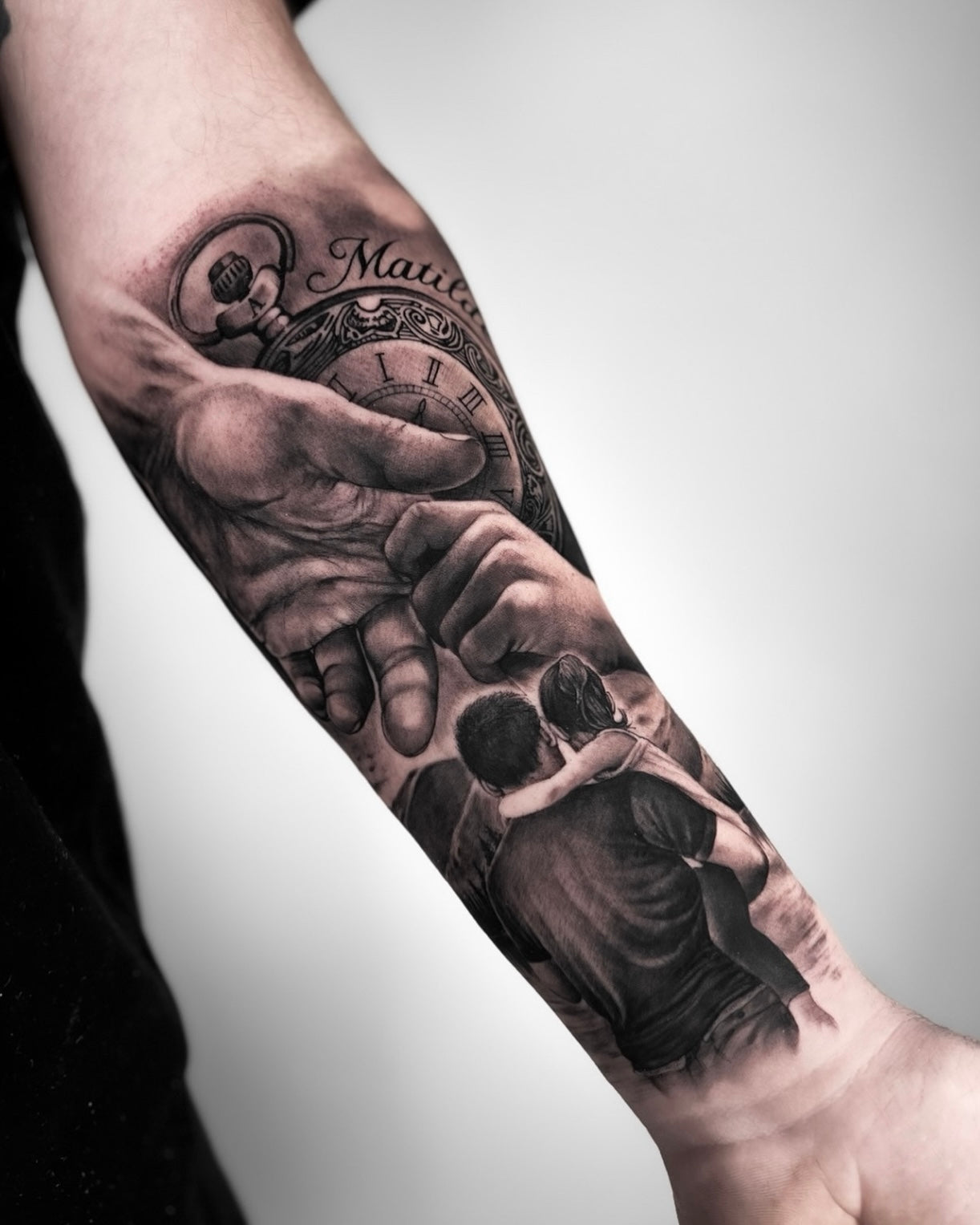 Tatuagem realista em black and grey de mão a segurar relógio de bolso e mão de criança com lettering personalizado no antebraço, tema familiar, Dilúvio Tattoo Benfica.
