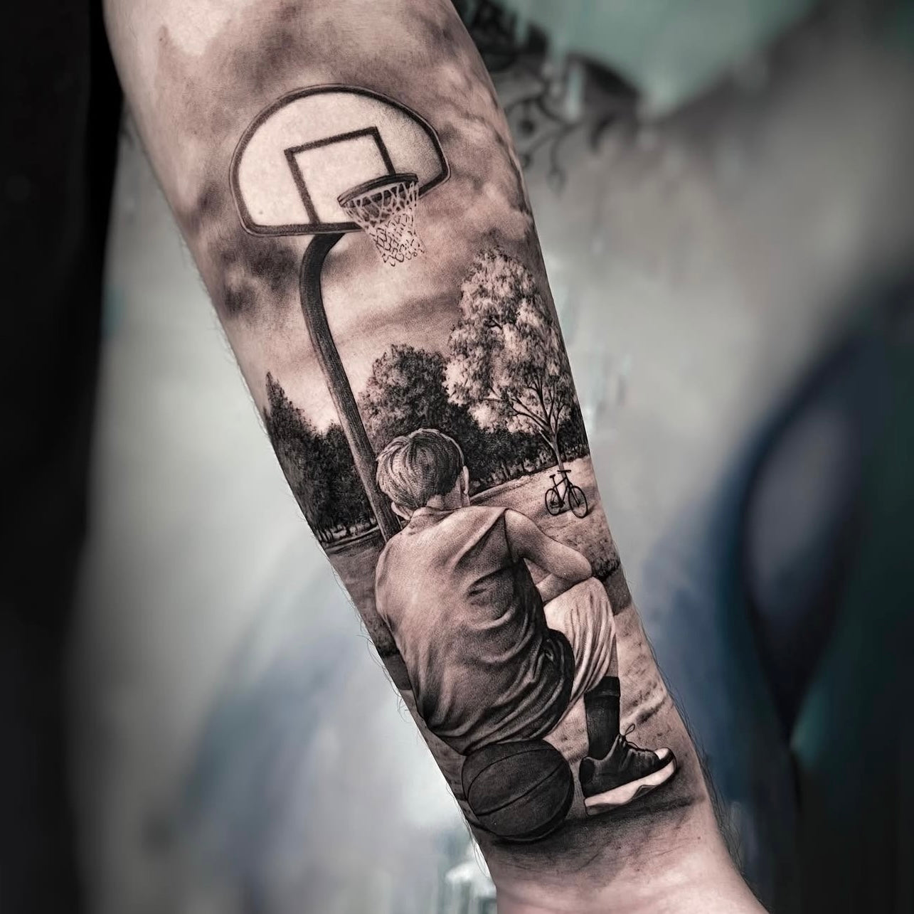 atuagem realista em black and grey de cesto de basquetebol com criança sentada e bola no antebraço, estilo cena urbana, Dilúvio Tattoo Benfica.