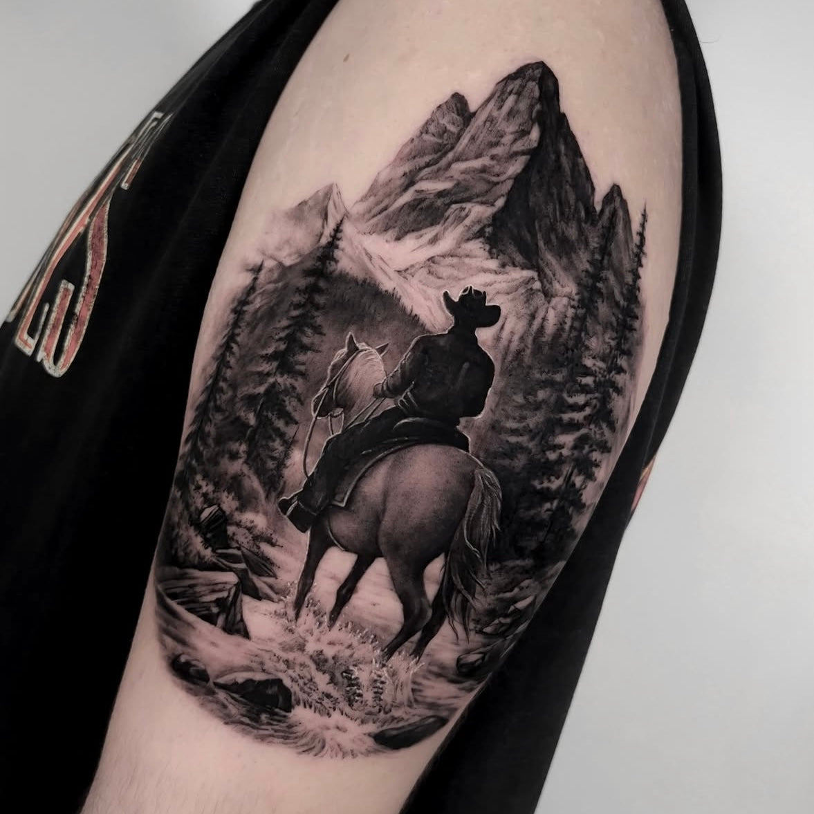 Tatuagem realista em black and grey de cowboy a cavalo numa paisagem com montanhas e floresta no braço, Dilúvio Tattoo Benfica.
