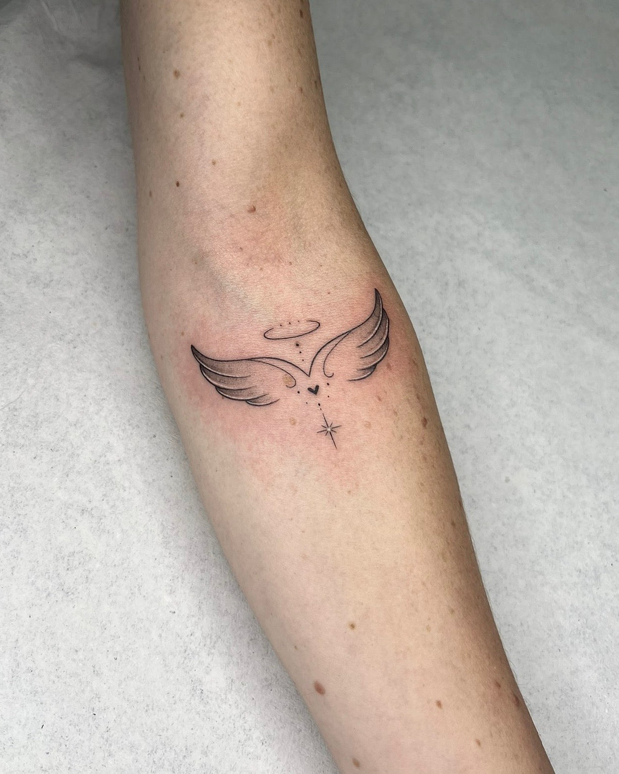 Tatuagem fine line de asas de anjo com halo e pequenas estrelas no braço, estilo dedicatória delicada, no Dilúvio Tattoo em Benfica.