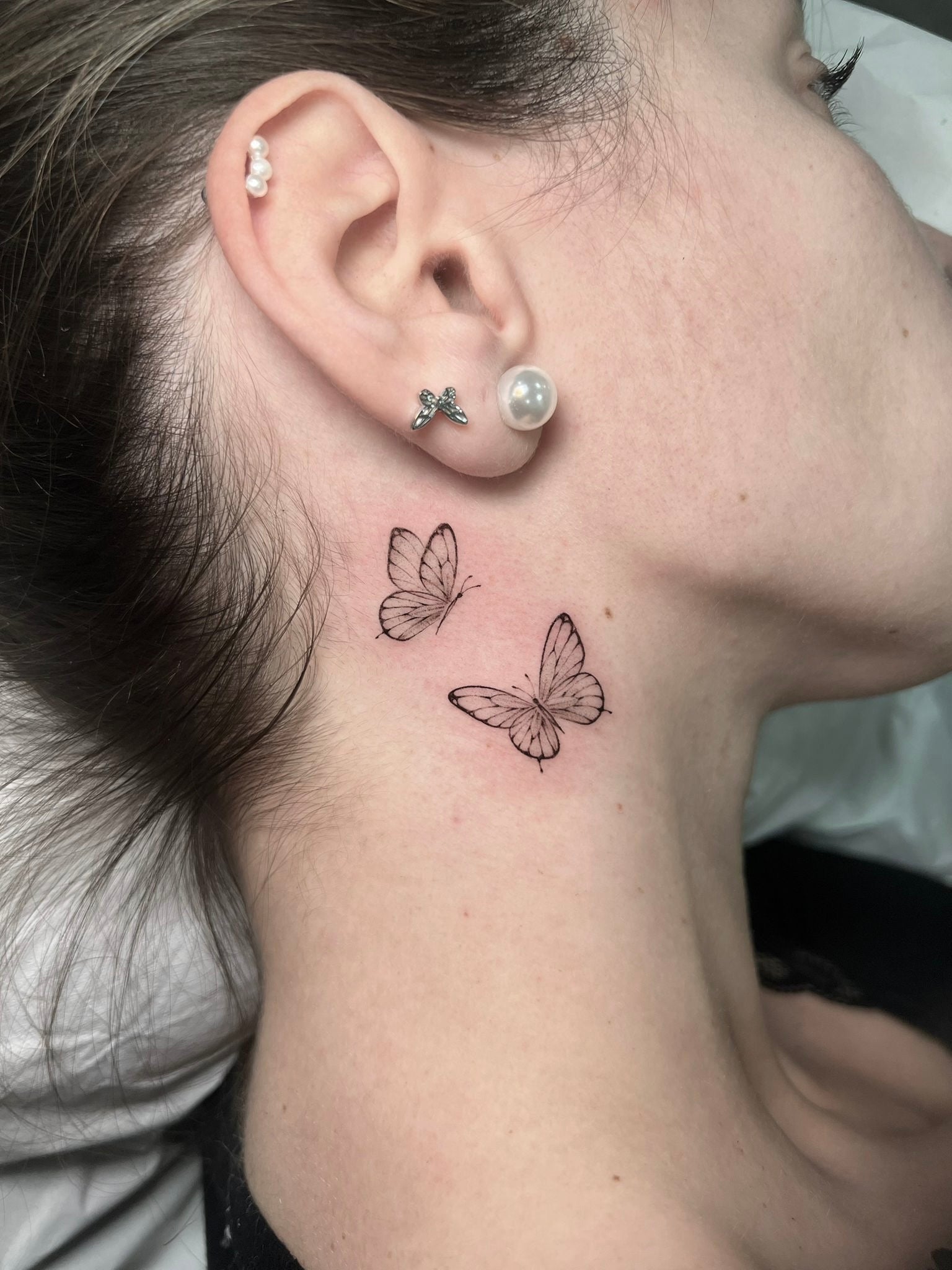 Tatuagem fine line de duas borboletas pequenas atrás da orelha no pescoço, estilo delicado feminino, no Dilúvio Tattoo em Benfica.