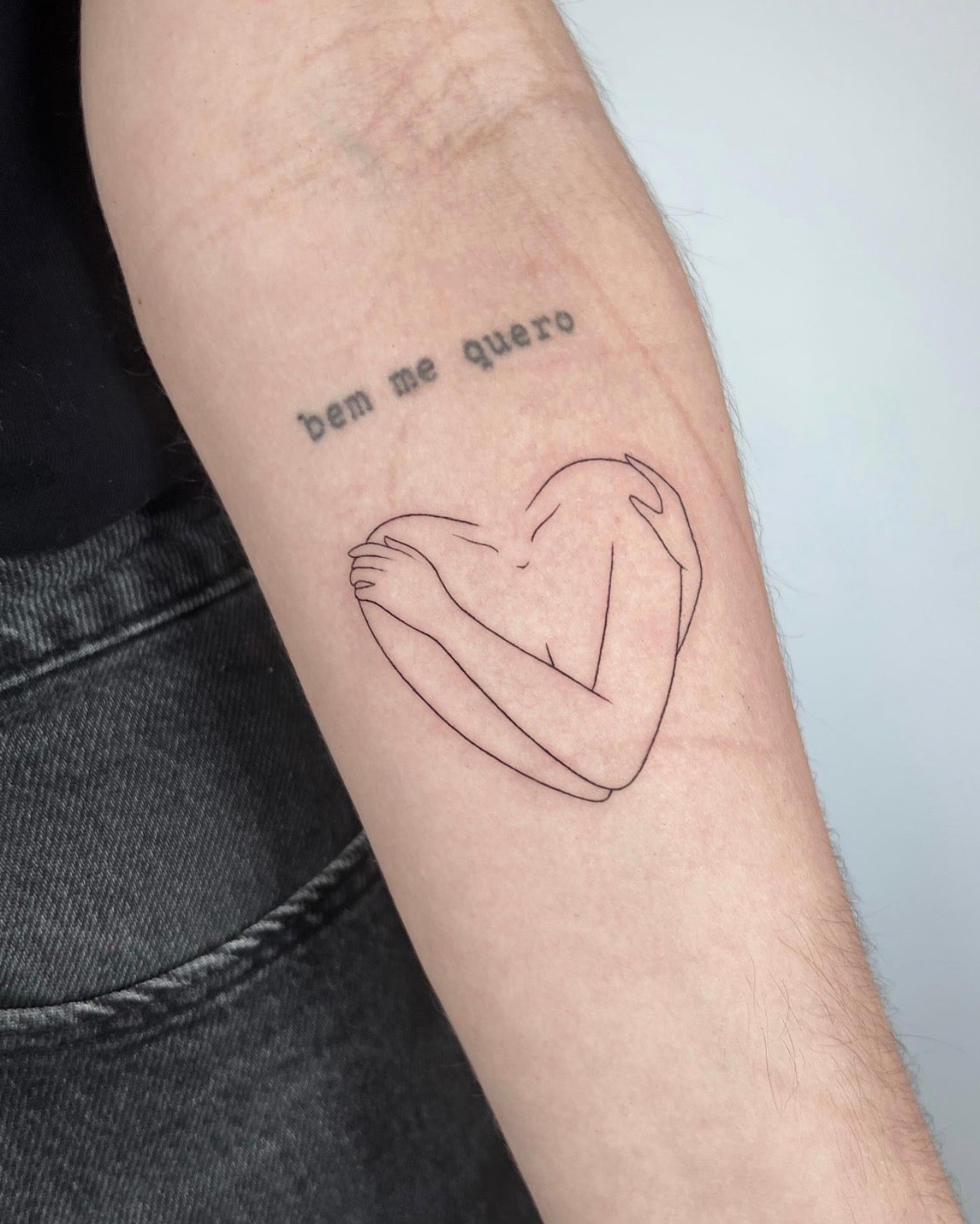 Tatuagem fine line de coração abraçado por braços no antebraço, estilo minimalista, feita no Dilúvio Tattoo em Benfica, Lisboa