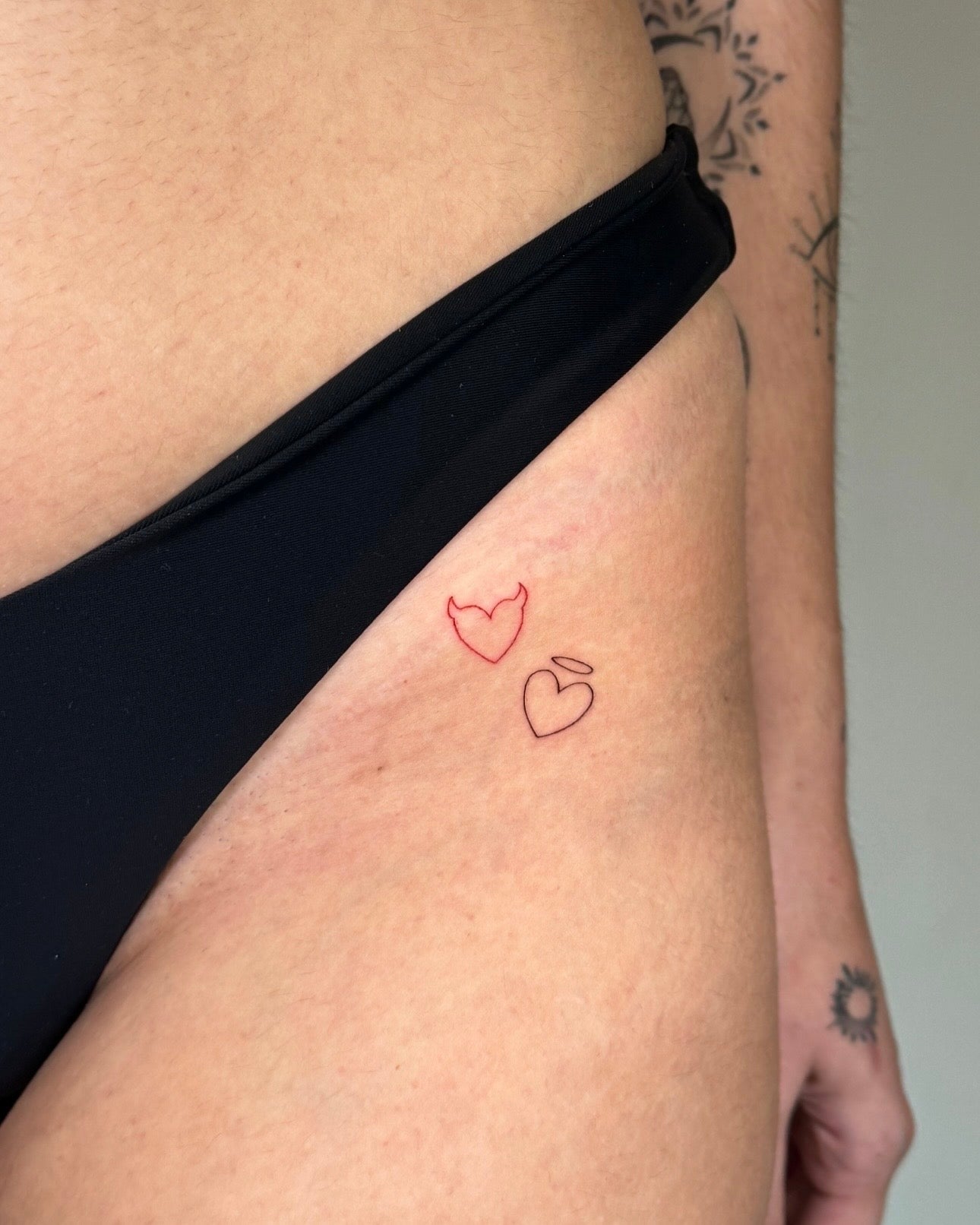 Tatuagem fine line minimalista de dois corações pequenos, um a vermelho e outro a preto, na anca, no estúdio Dilúvio em Benfica, Lisboa.