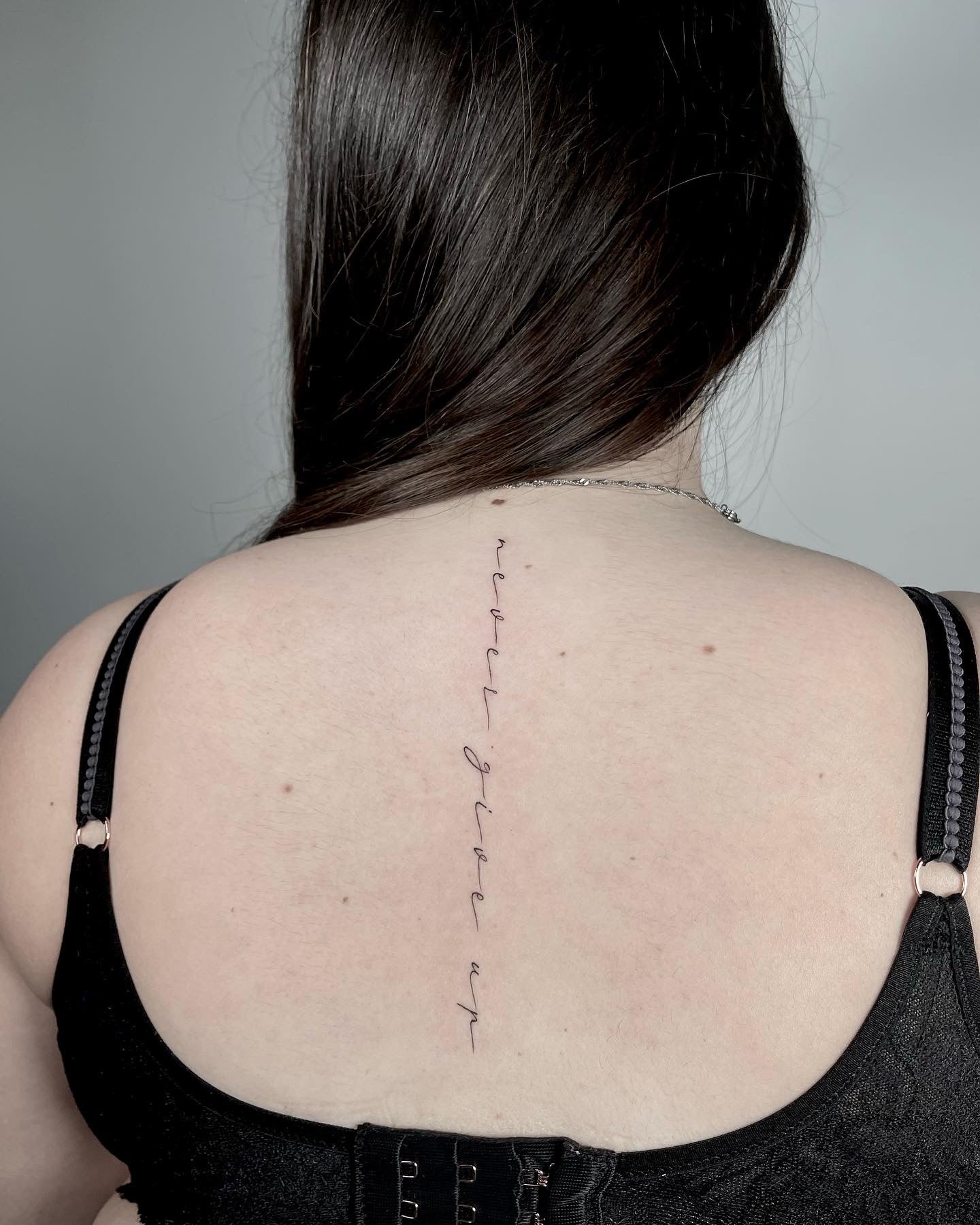 Tatuagem fine line de frase em lettering cursivo delicado ao longo da coluna nas costas, estilo caligráfico feminino, no Dilúvio Tattoo em Benfica.