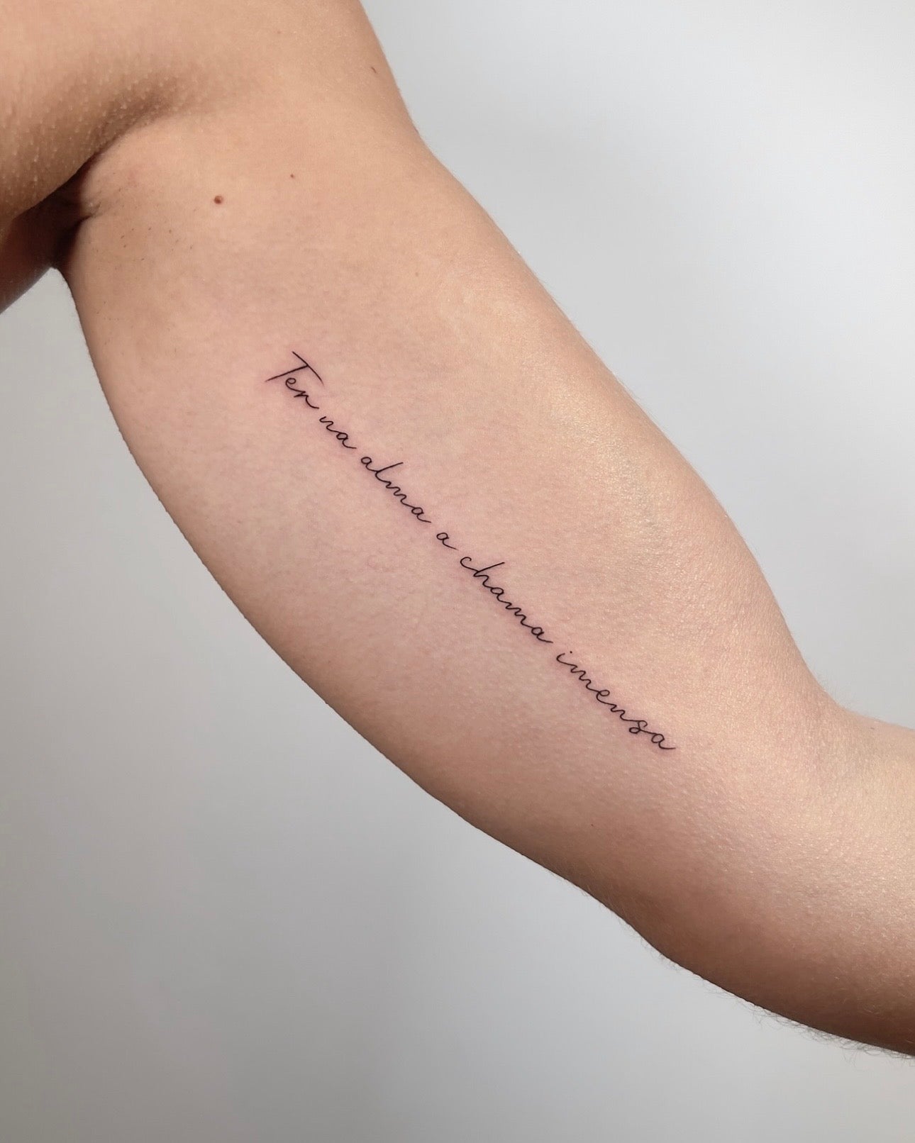 Tatuagem fine line de frase personalizada em lettering cursivo diagonal no braço, estilo caligráfico minimalista, no Dilúvio Tattoo em Benfica, Lisboa.