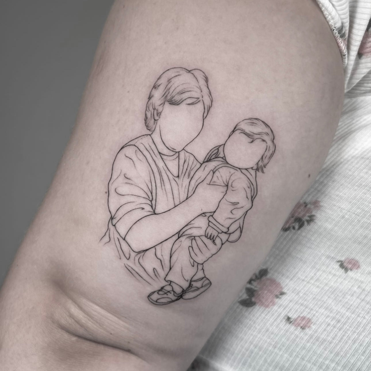 Tatuagem fine line de figura a segurar bebé ao colo, estilo retrato ilustrativo minimalista, dedicatória familiar, no Dilúvio Tattoo em Benfica.