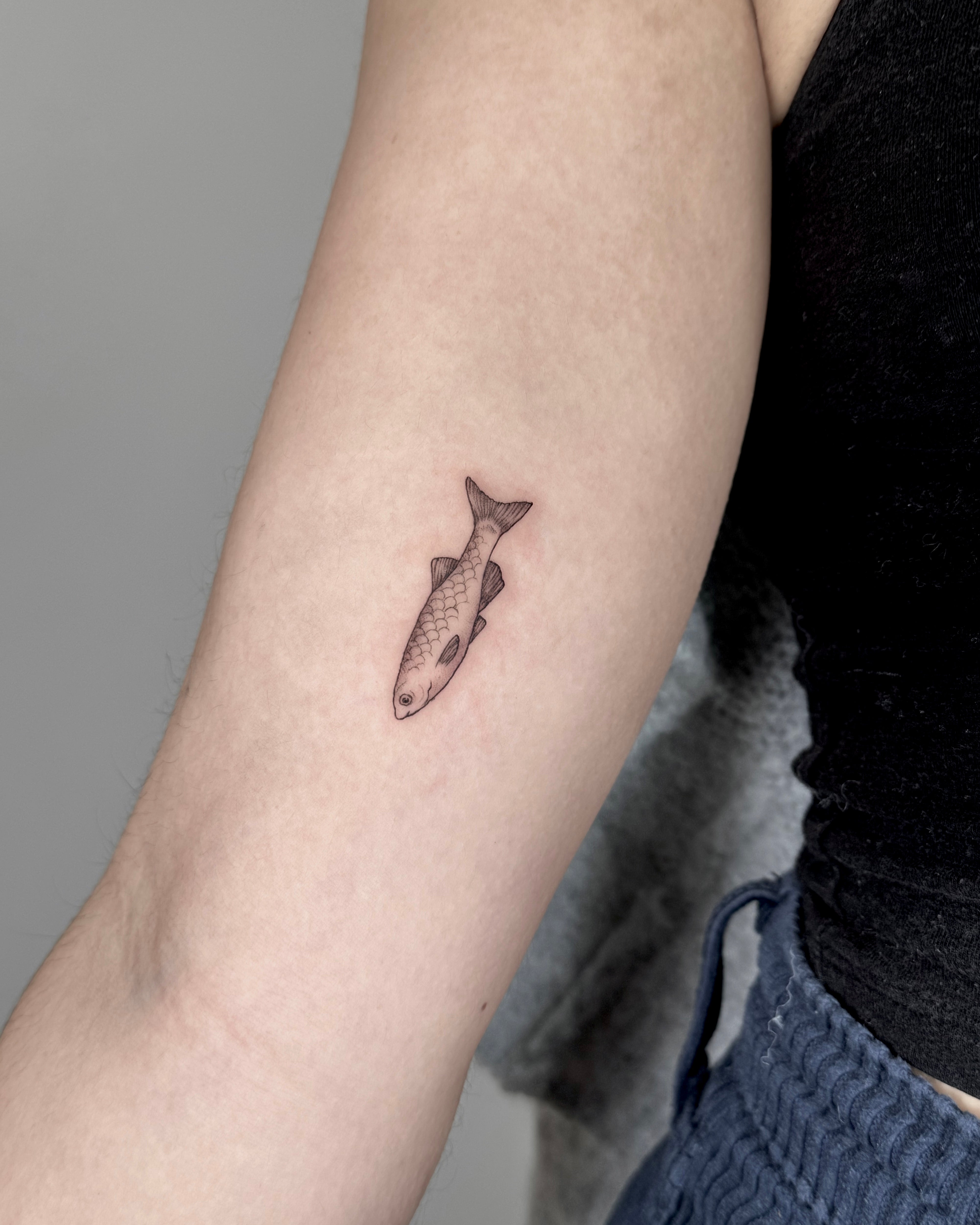 Tatuagem fine line de peixe pequeno com detalhes de escamas no braço, estilo minimalista, no Dilúvio Tattoo em Benfica, Lisboa.