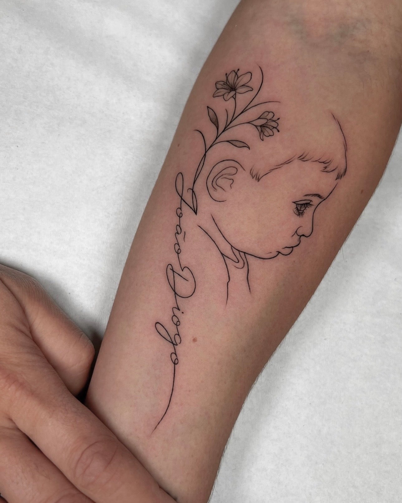 Tatuagem fine line de perfil de bebé com flor e lettering do nome 