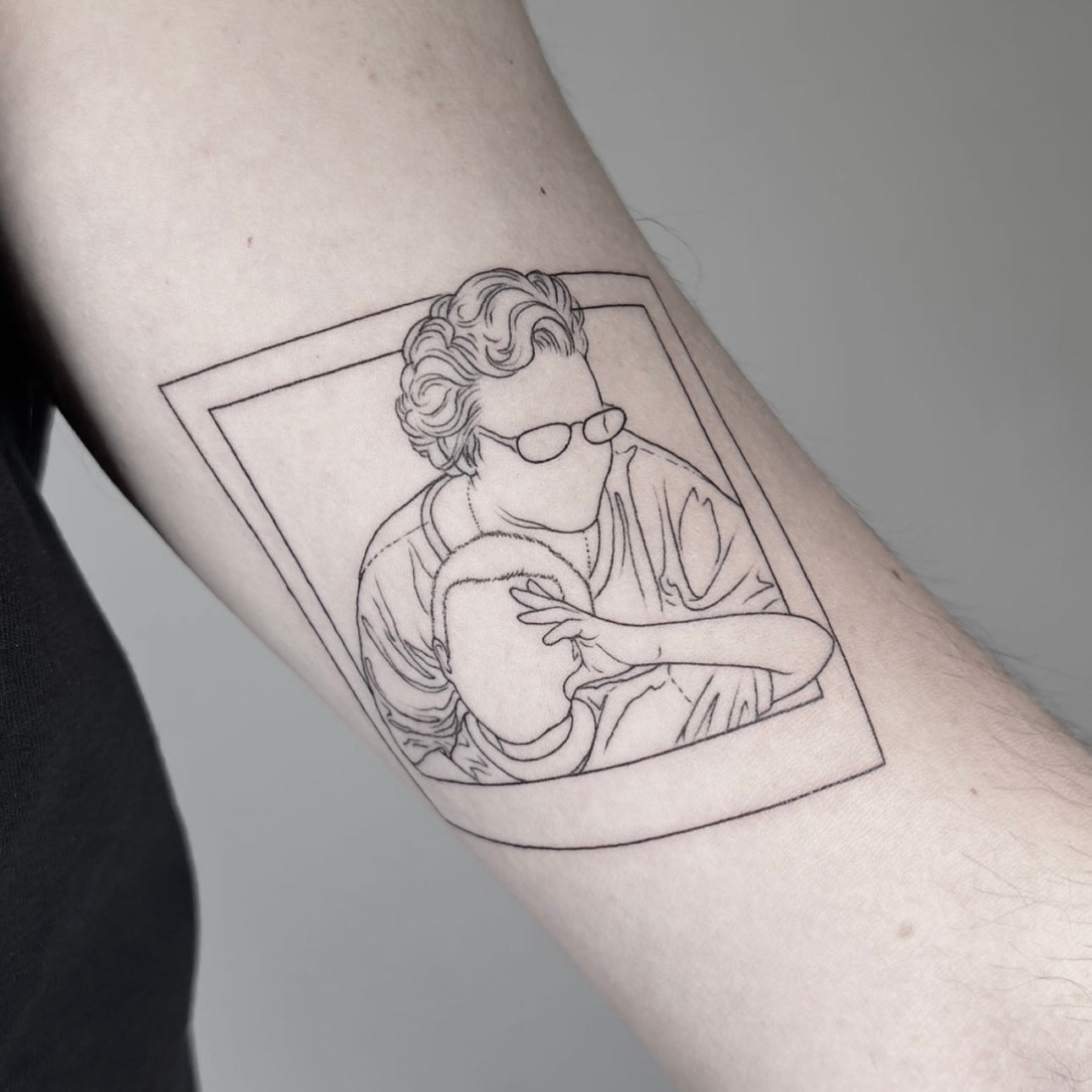 Tatuagem fine line de retrato ilustrado dentro de moldura tipo polaroid a segurar um bebé no braço, estilo memória familiar, no Dilúvio Tattoo em Benfica.