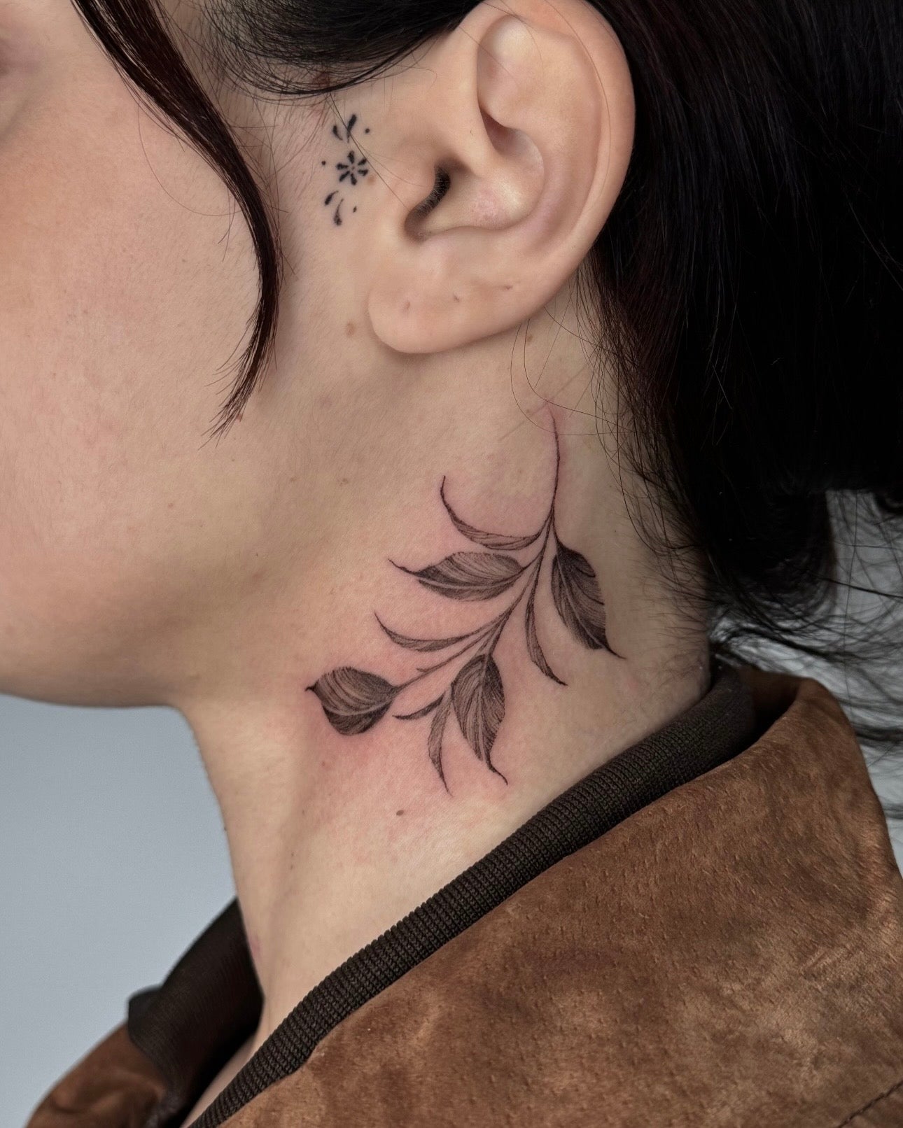 Tatuagem fine line feminina de ramo de folhas no pescoço com pequenas flores atrás da orelha, estilo delicado botânico, no Dilúvio Tattoo Benfica.