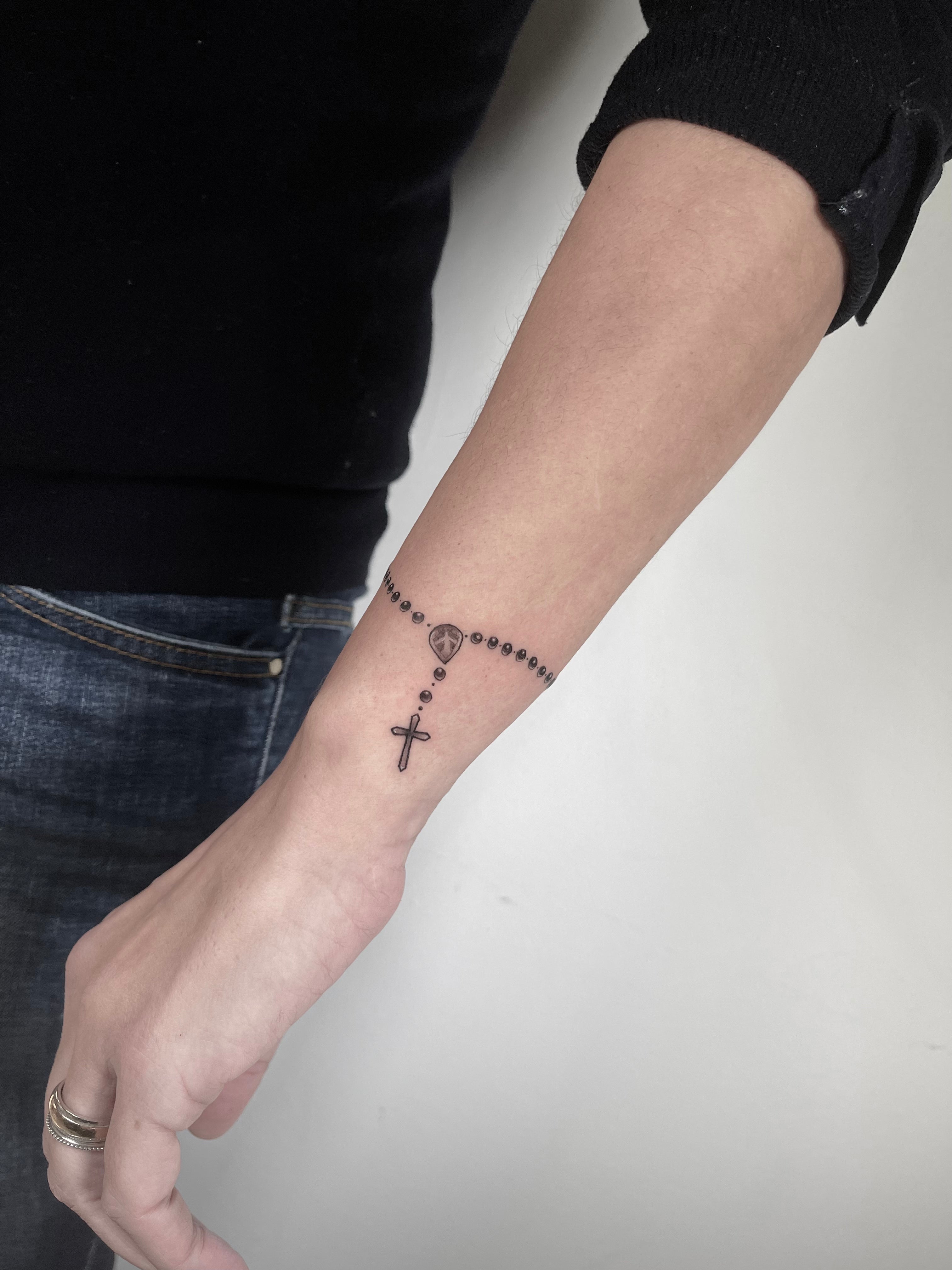 Tatuagem fine line de terço com contas e cruz a envolver o pulso, estilo religioso delicado, no Dilúvio Tattoo em Benfica, Lisboa.