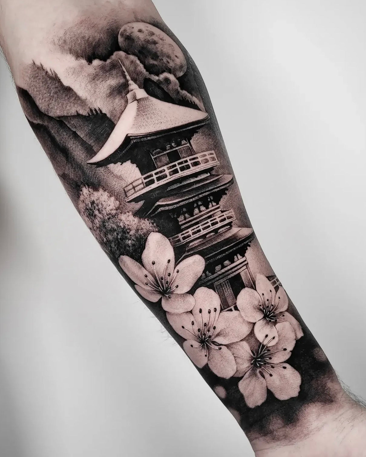 Tatuagem em black and grey japonês rodeado por flores de cerejeira e montanhas no antebraço, estilo realismo oriental, Dilúvio Tattoo Benfica.