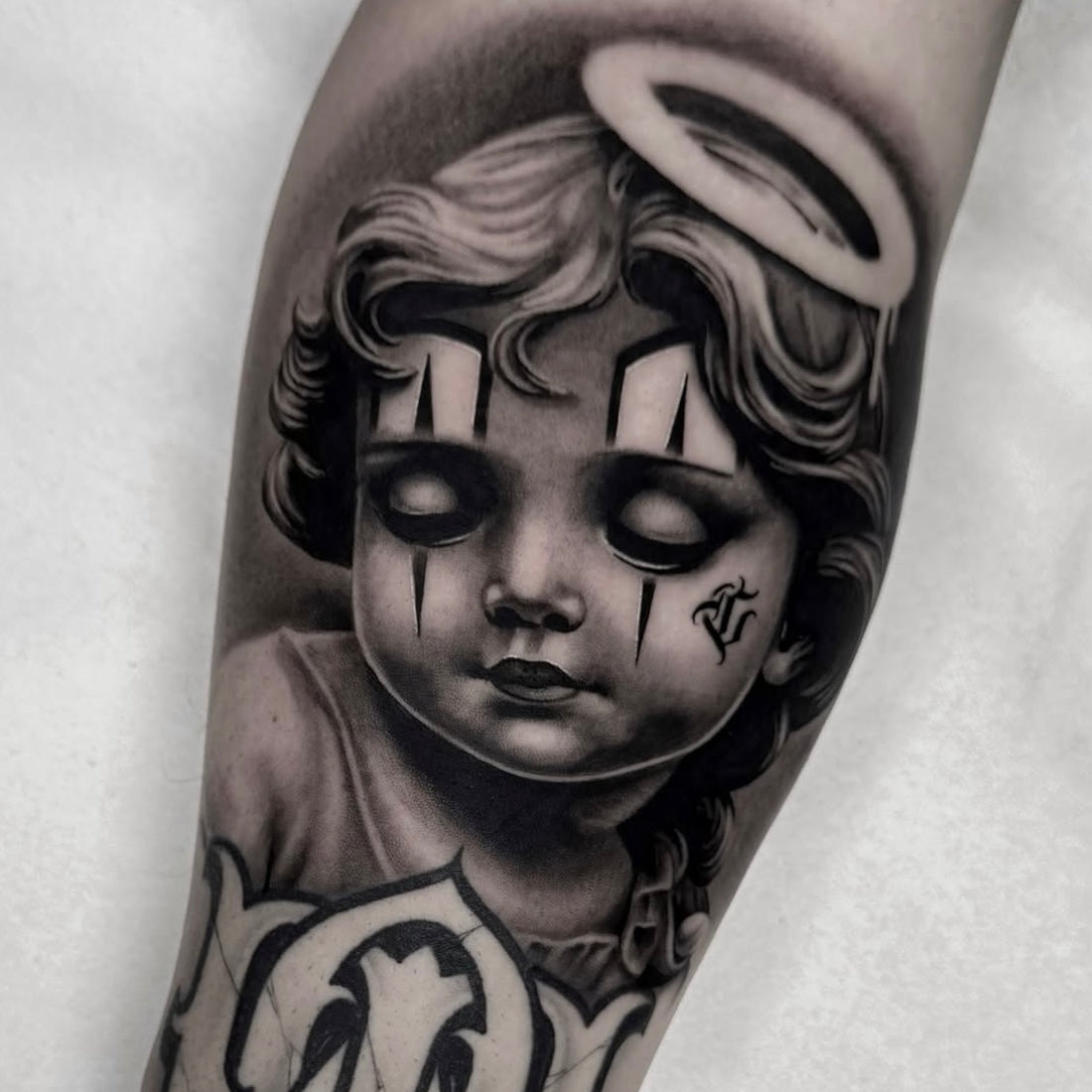 Tatuagem em black and grey de querubim com halo e lágrimas de palhaço no antebraço, inspiração chicano/religioso, feita no Dilúvio Tattoo em Benfica
