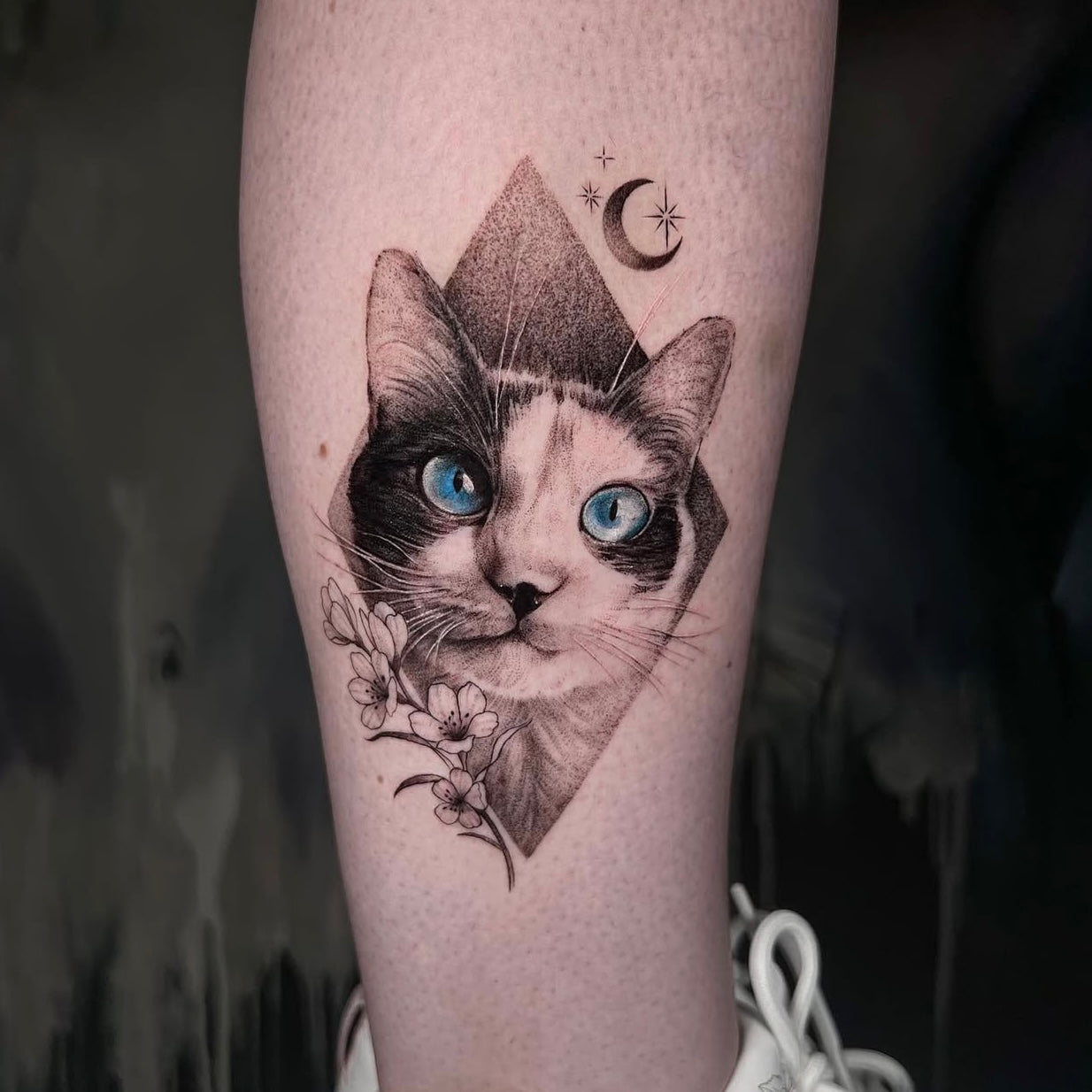 Tatuagem realista de retrato de gato siamês com olhos azuis dentro de losango com lua e flores no braço, black and grey com pop of color, homenagem a animal de estimação, na Dilúvio Tattoo em Benfica.