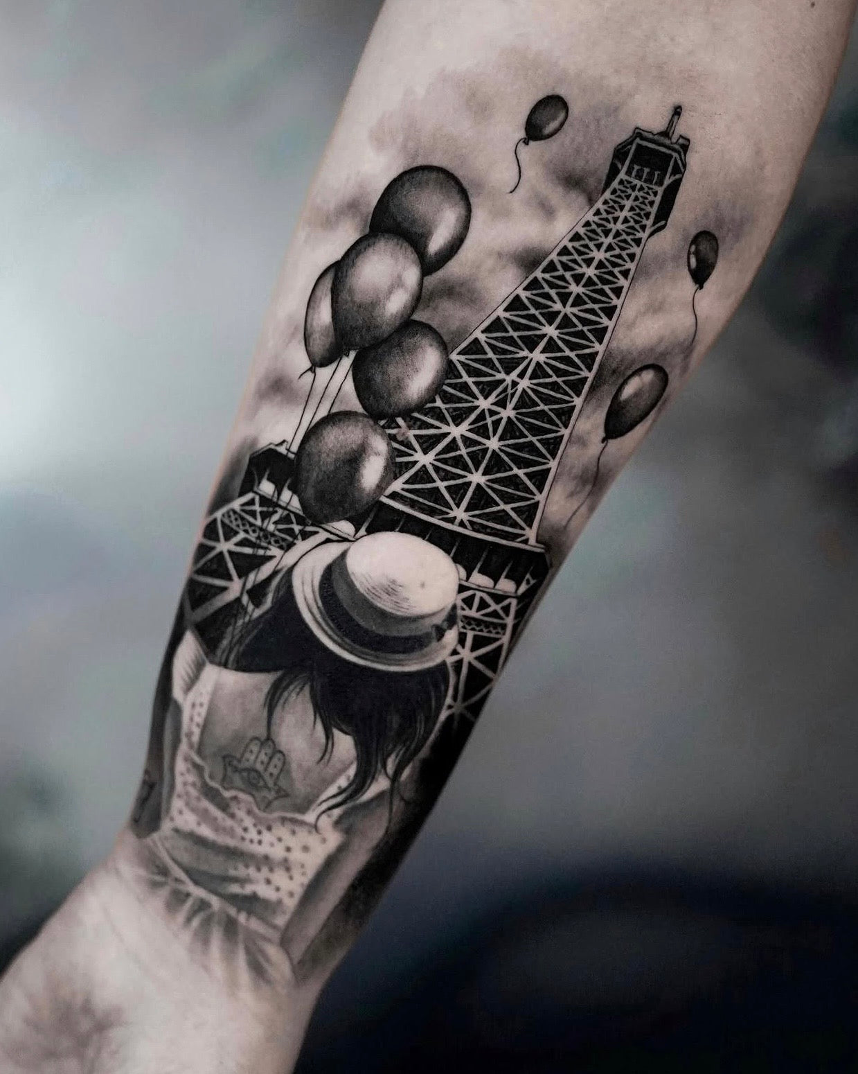 Tatuagem realista em black and grey de Torre Eiffel com balões e menina de chapéu de palha no braço, estilo cinemático, feita na Dilúvio Tattoo em Benfica, Lisboa.
